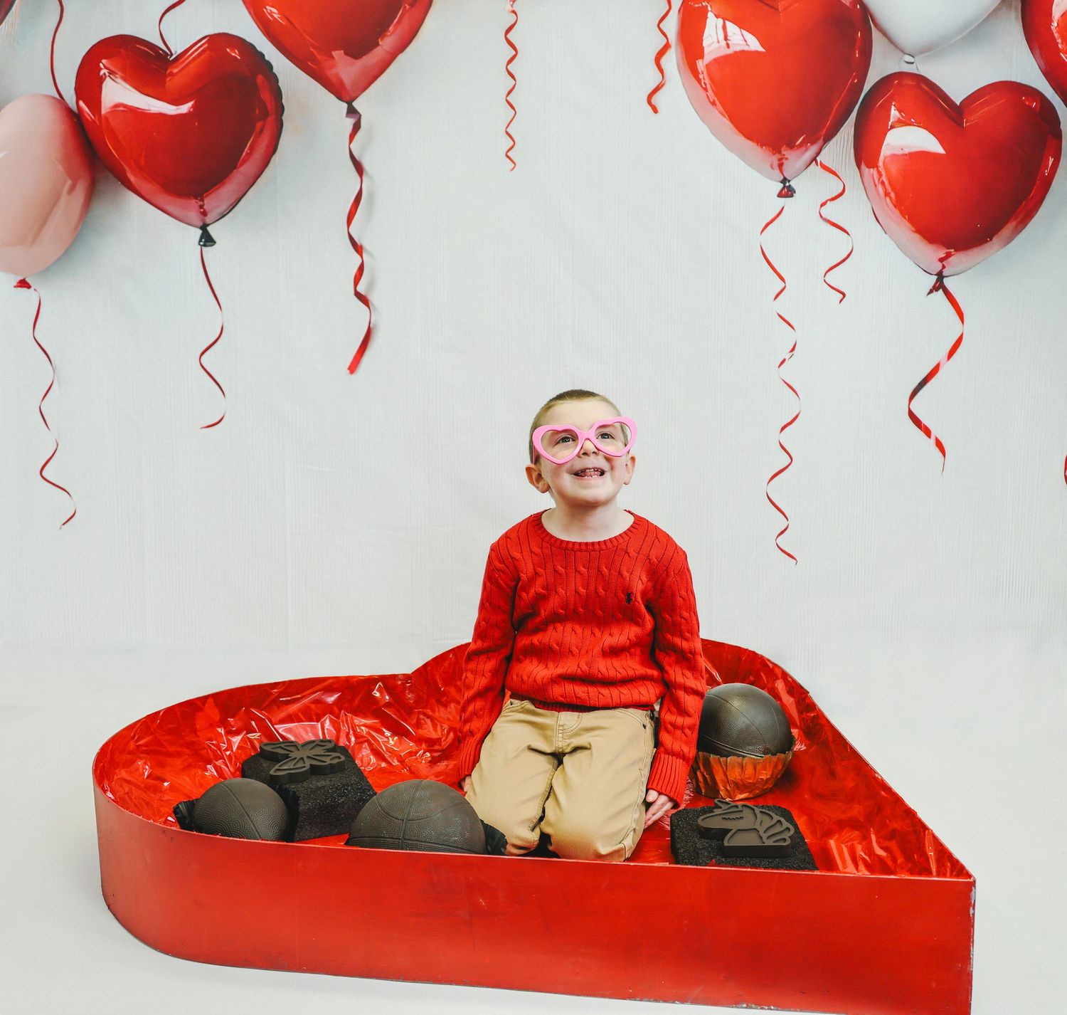 Smiling child in red sweater sitting in heart-shaped box with heart balloons, wearing pink glasses for Valentine's Day.
