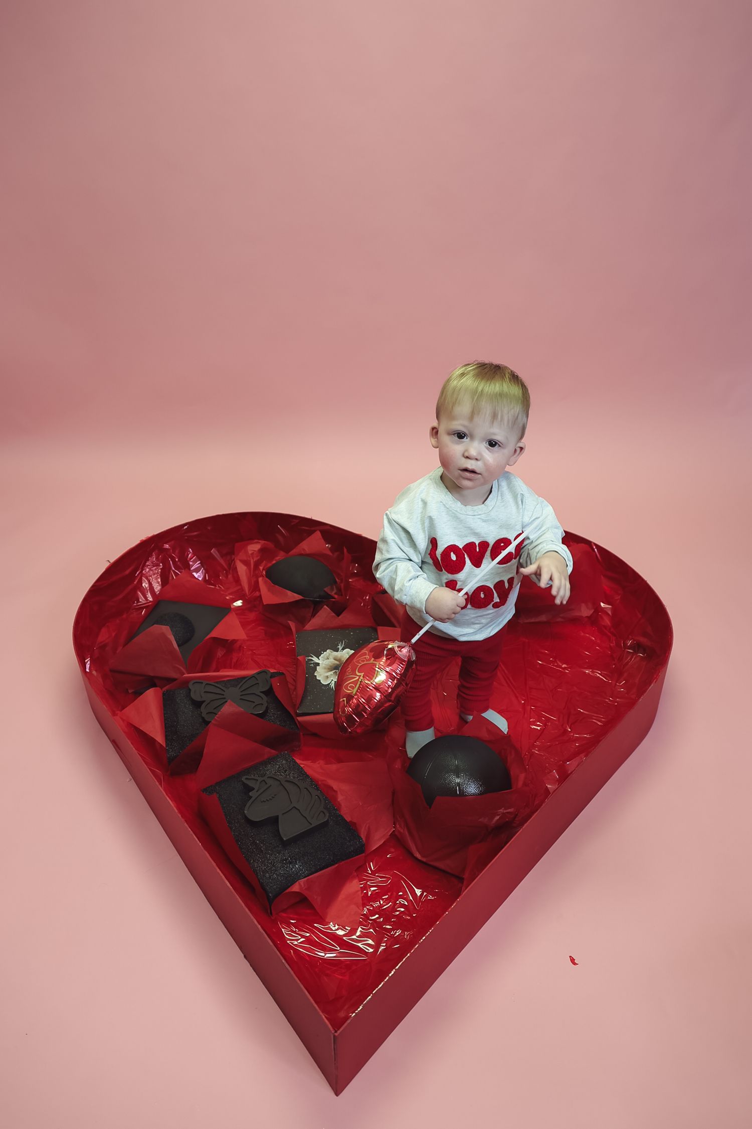 A baby sits inside a large red heart-shaped Valentine's Day chocolate box on a pink background.