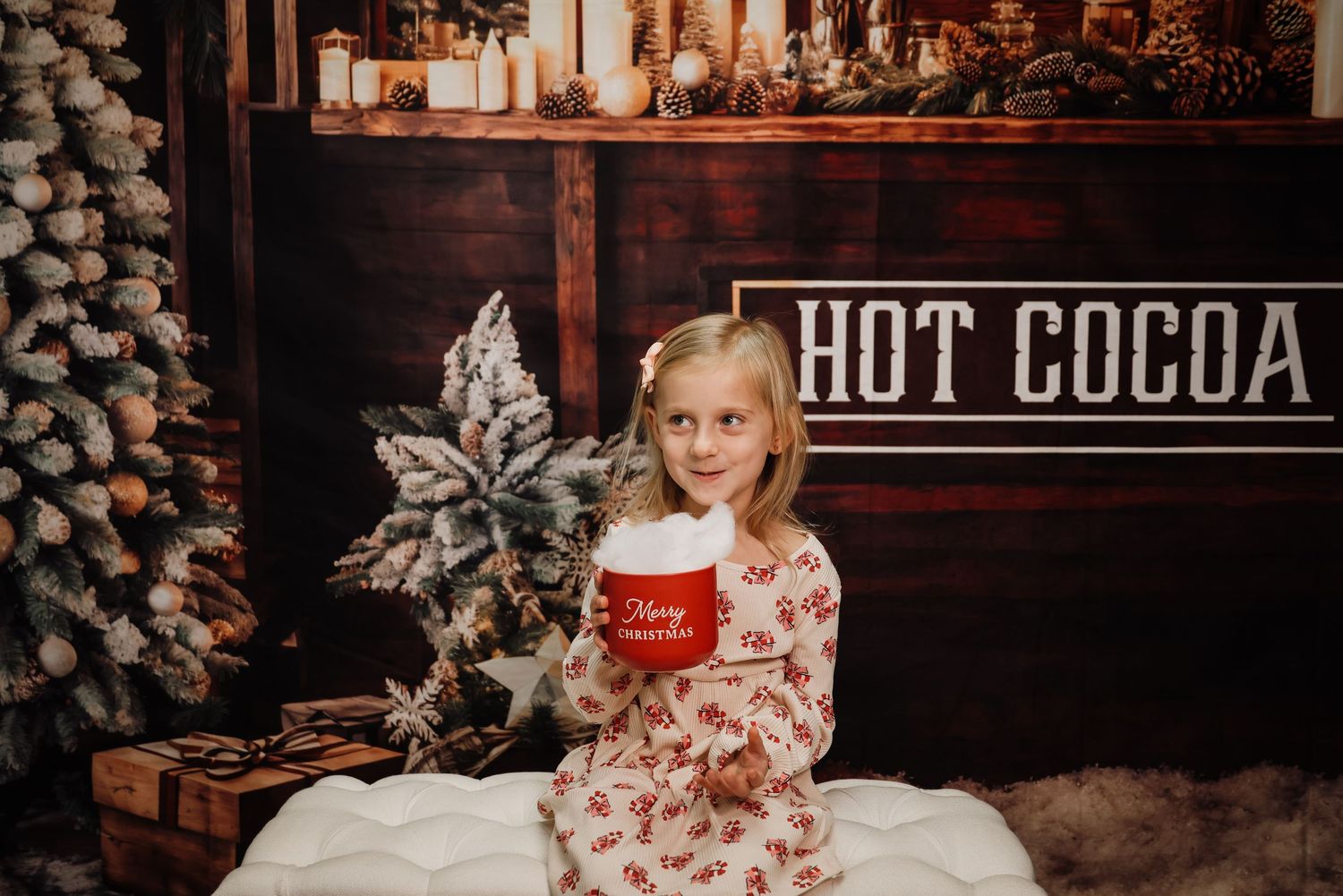 Young girl in holiday pajamas holds red hot cocoa mug, sitting near snowy Christmas tree with festive backdrop.