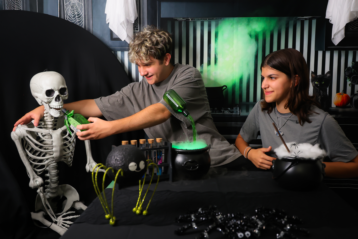 Two people decorating a Halloween table with a skeleton, bubbling cauldron, and spooky props.
