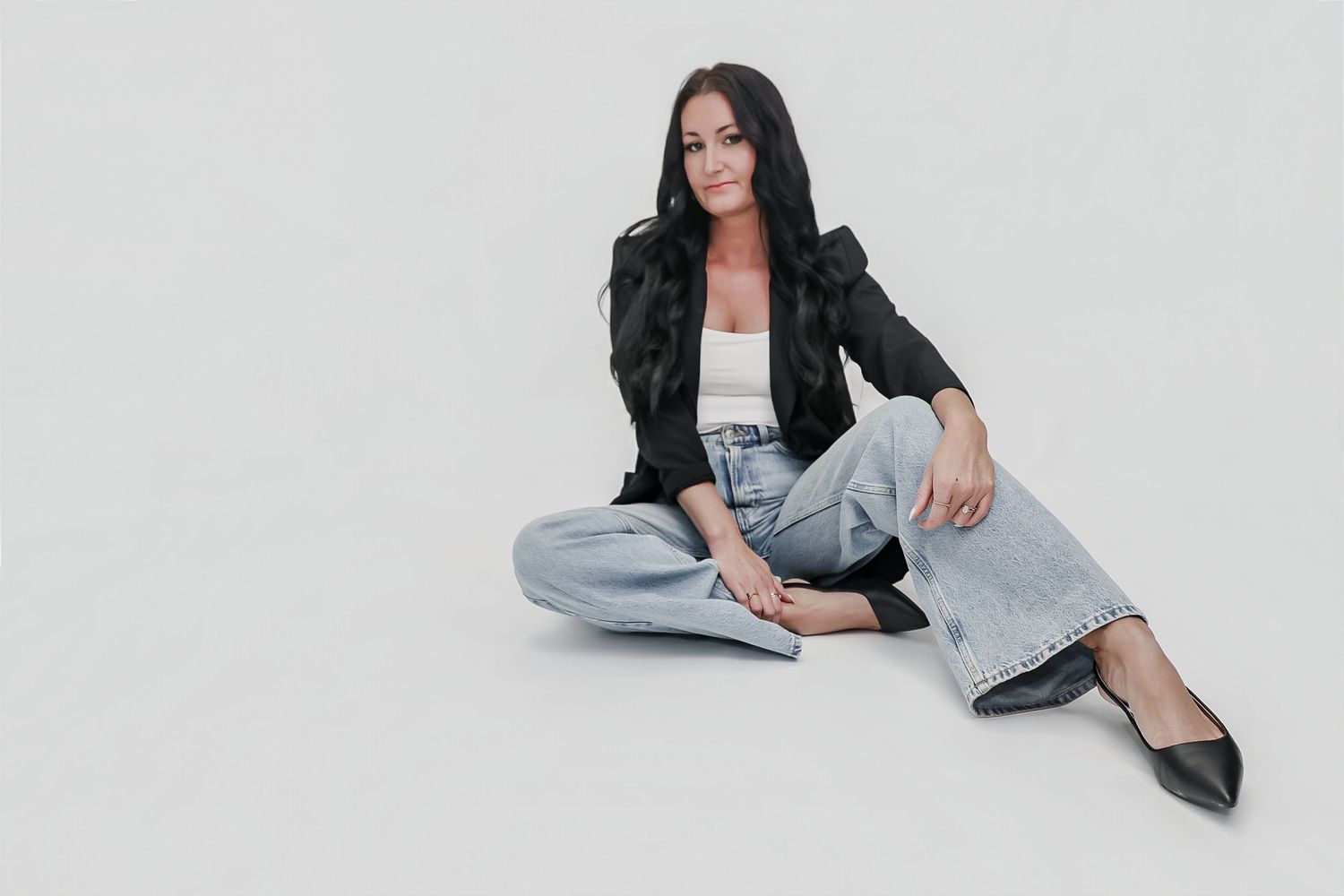Woman in black blazer, white top, and light jeans sits cross-legged on white floor, posing confidently.