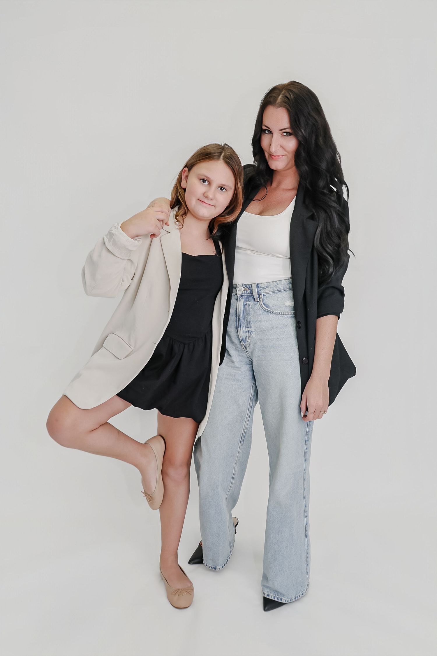 A woman and young girl pose together in stylish outfits, wearing blazers against a white background.