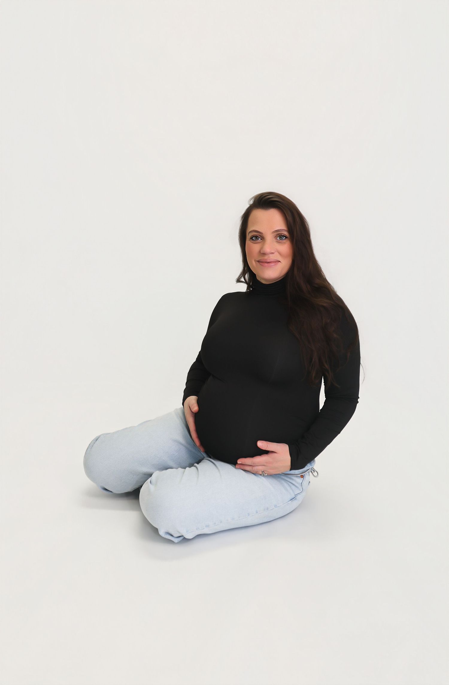 Pregnant woman in black turtleneck and light jeans sitting cross-legged, cradling her baby bump, smiling at camera.