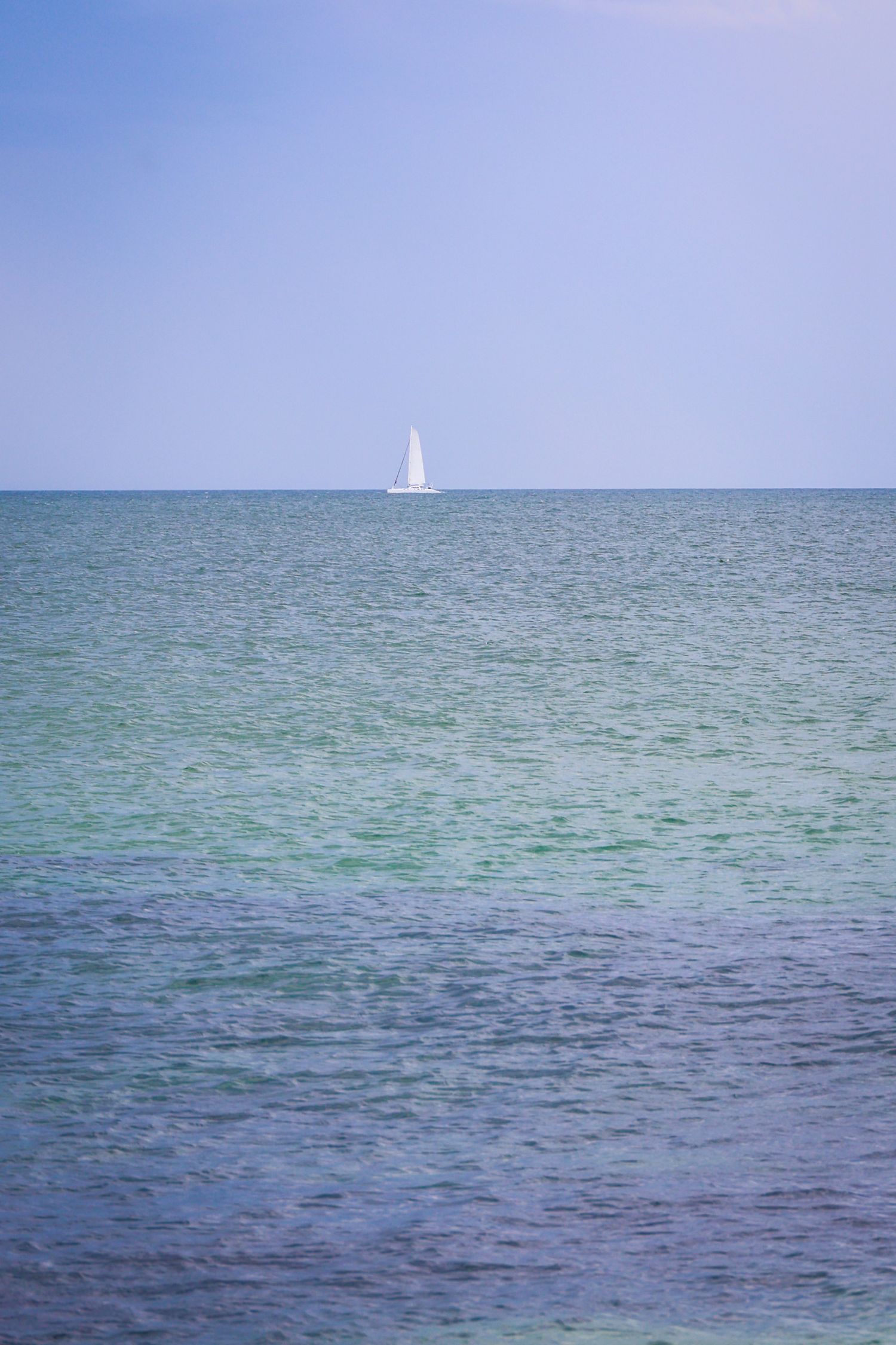 A white sailboat glides across calm turquoise ocean waters under a clear blue sky on a sunny day.