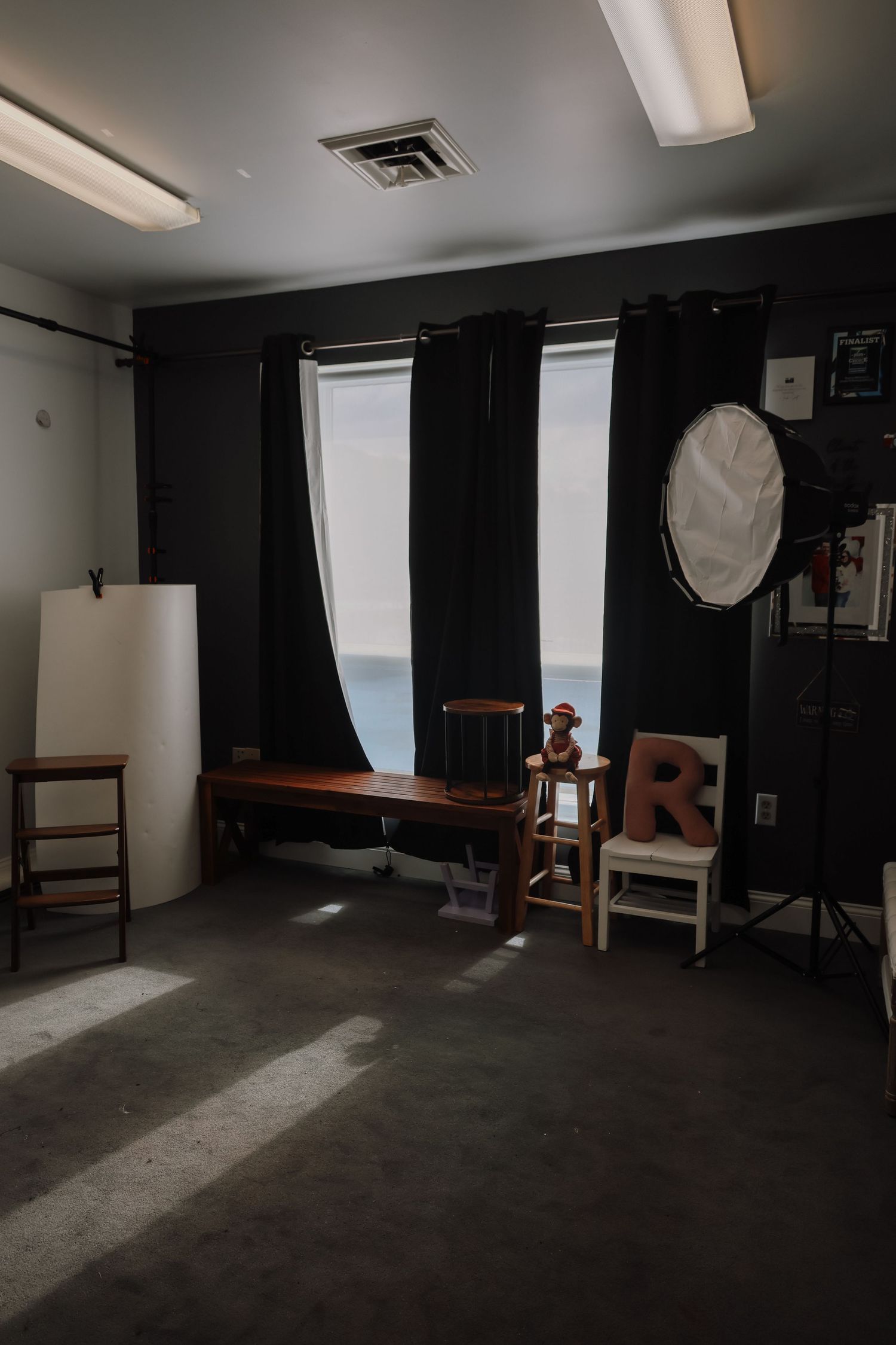 A dark photography studio interior with wooden chairs and softbox lighting equipment against black walls and natural window light.