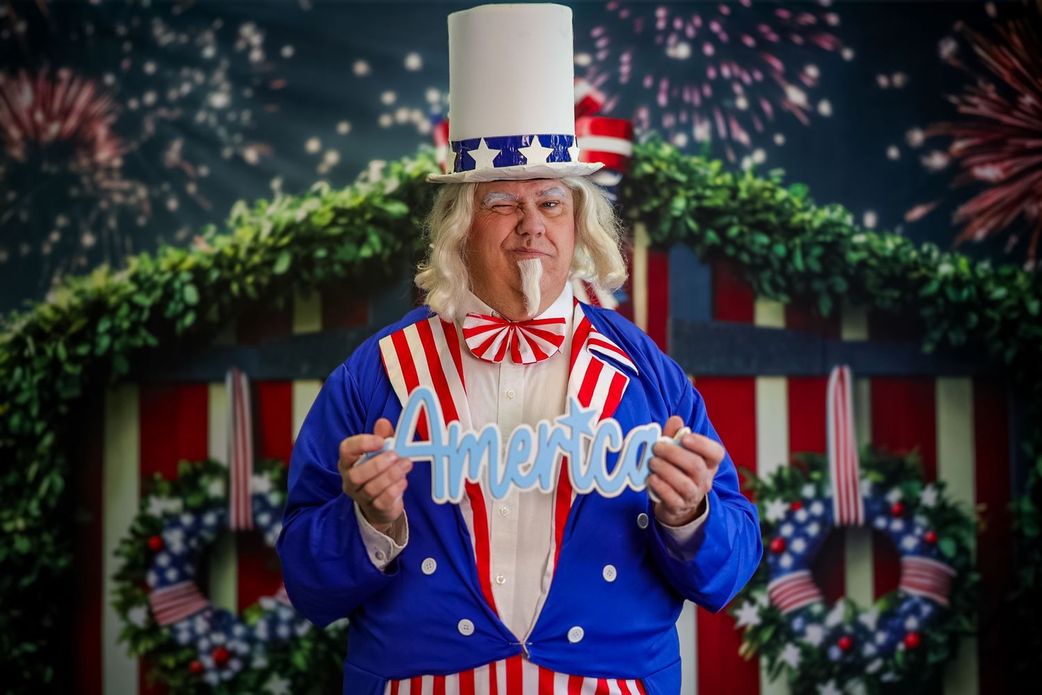 Person dressed as Uncle Sam in patriotic costume holds an America sign with fireworks backdrop.