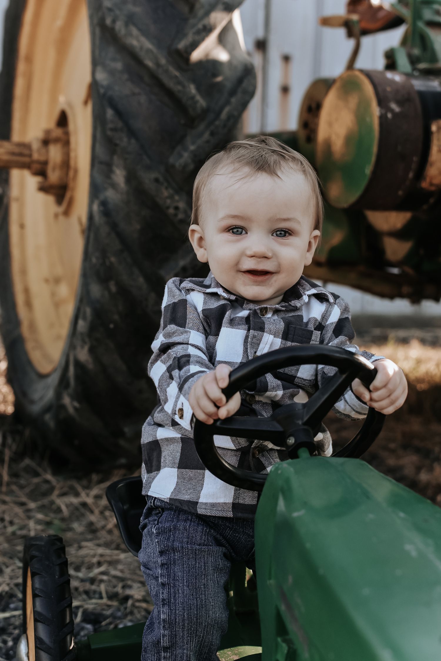 A baby in a plaid shirt and jeans smiles while sitting on a green tractor.