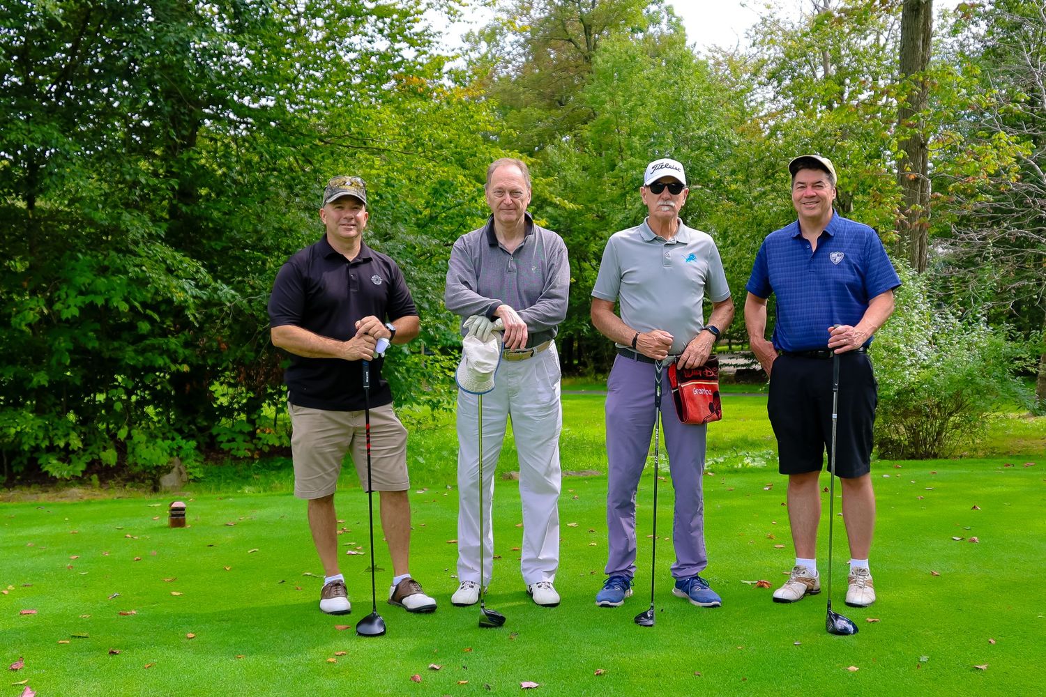 Four golfers stand together on a lush green golf course with clubs in hand, surrounded by trees and foliage.