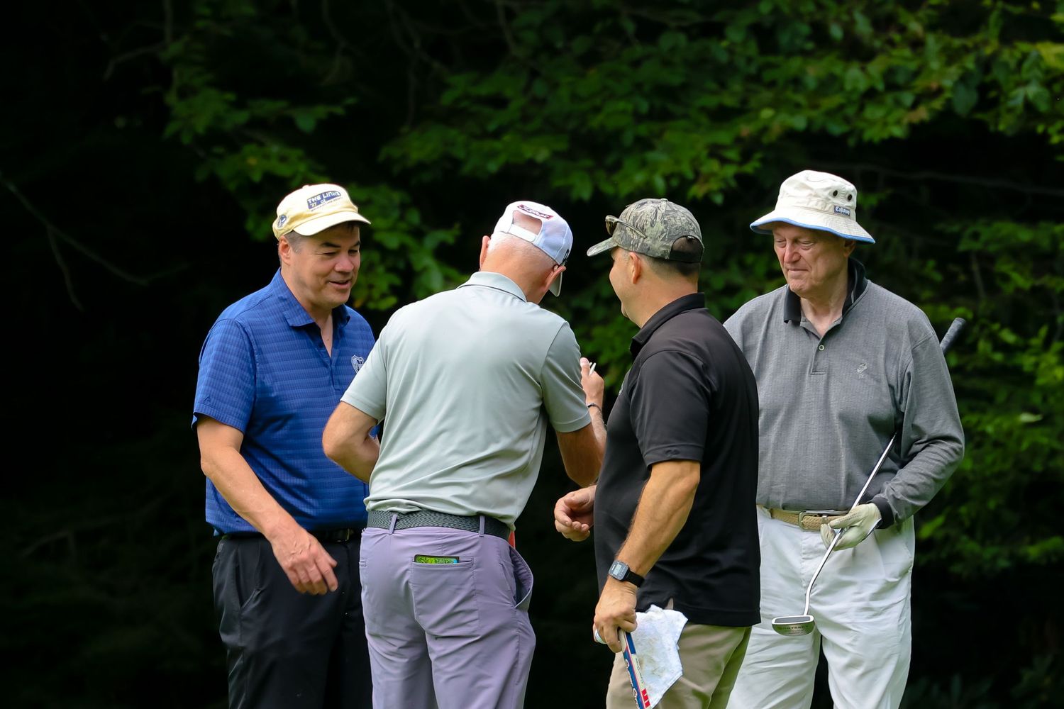 Golfers gather on a lush green course during a game, wearing casual attire and caps in the summer weather.
