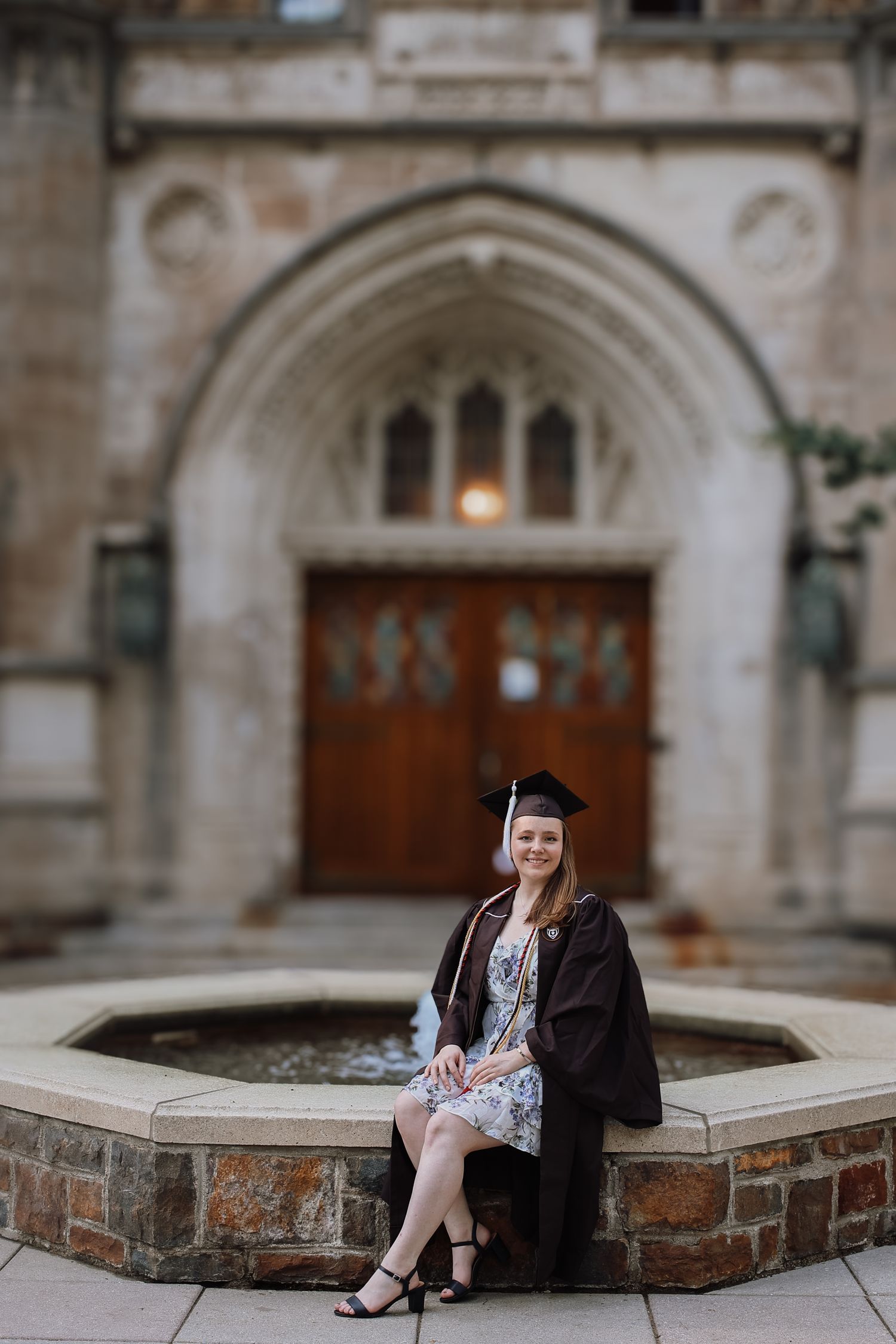 Graduate sits on stone fountain ledge in front of ornate gothic architecture wearing cap and gown for graduation photos.