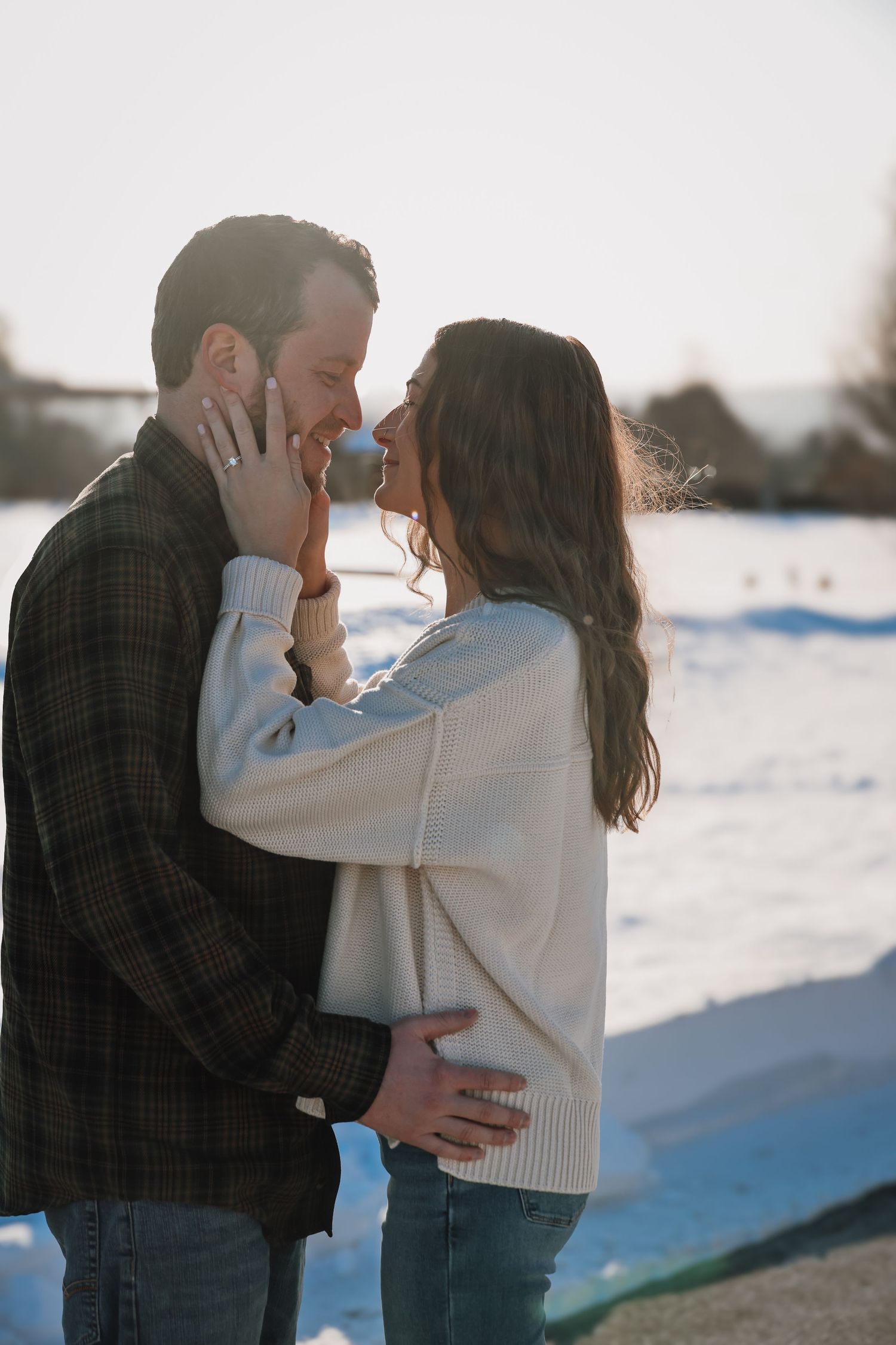 A romantic winter scene shows a couple embracing in the snow during golden hour, with soft sunlight illuminating their tender moment.