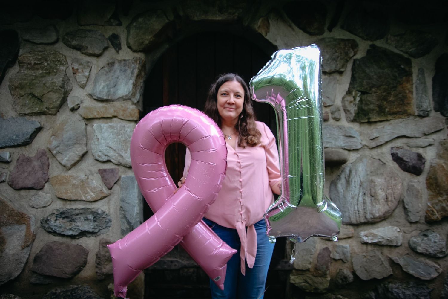 Someone stands against a stone wall holding large pink and green inflatable balloon decorations.