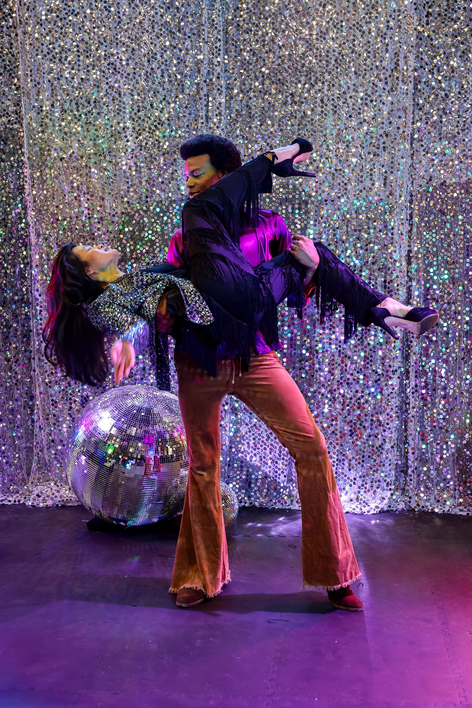 A dancer in flared pants performs a backbend sequence against a sparkly silver backdrop with a disco ball.