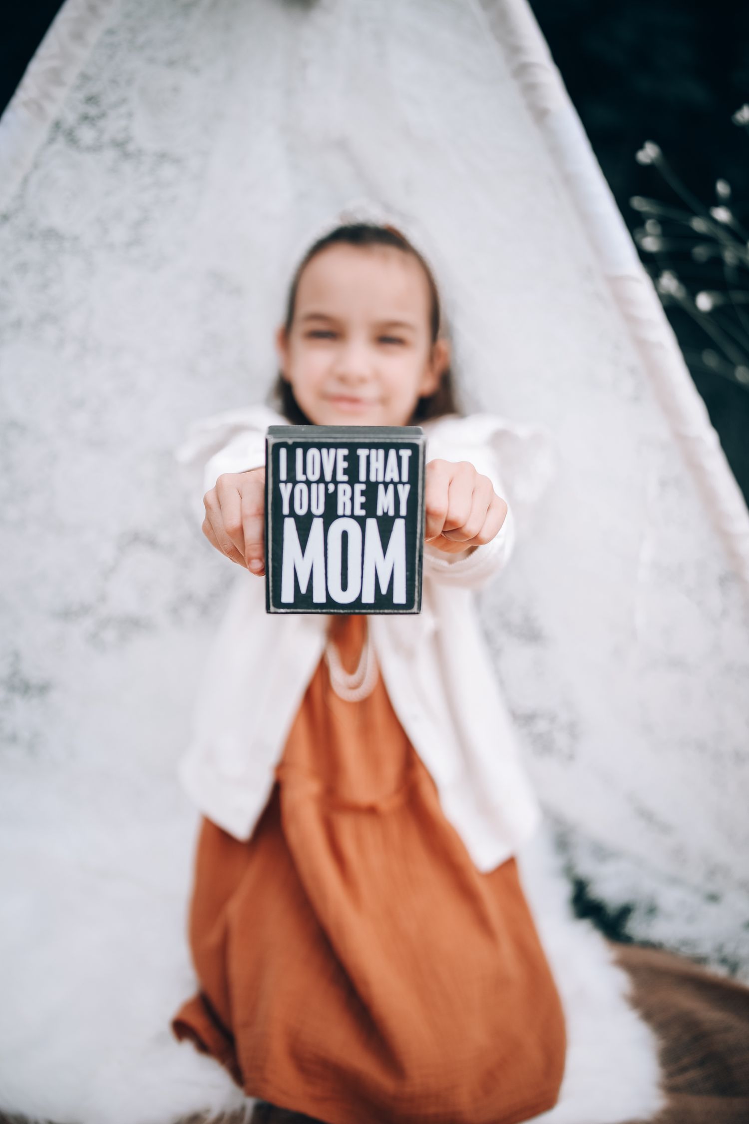 Someone in a white cardigan and rust-colored dress holds up a black sign reading 'I love that you're my mom' against a white wall.