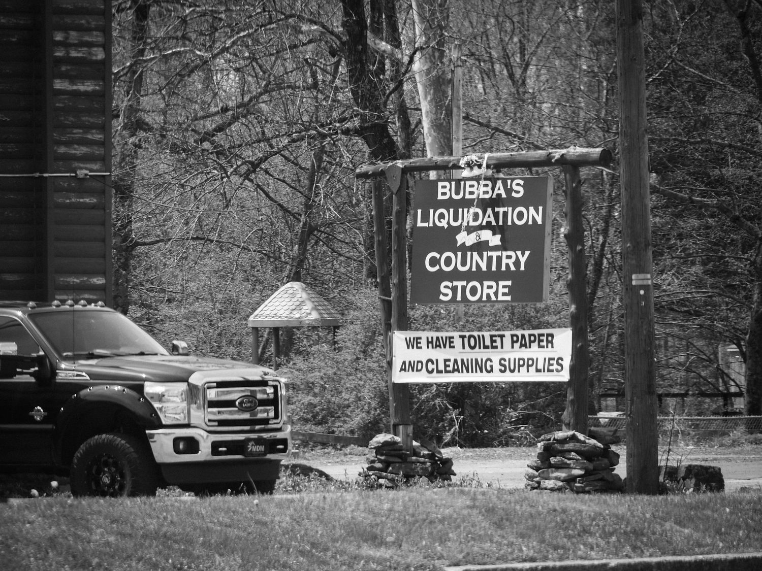 Black and white photo of Bubba's Location Country Store sign with pickup truck parked nearby in rural setting.