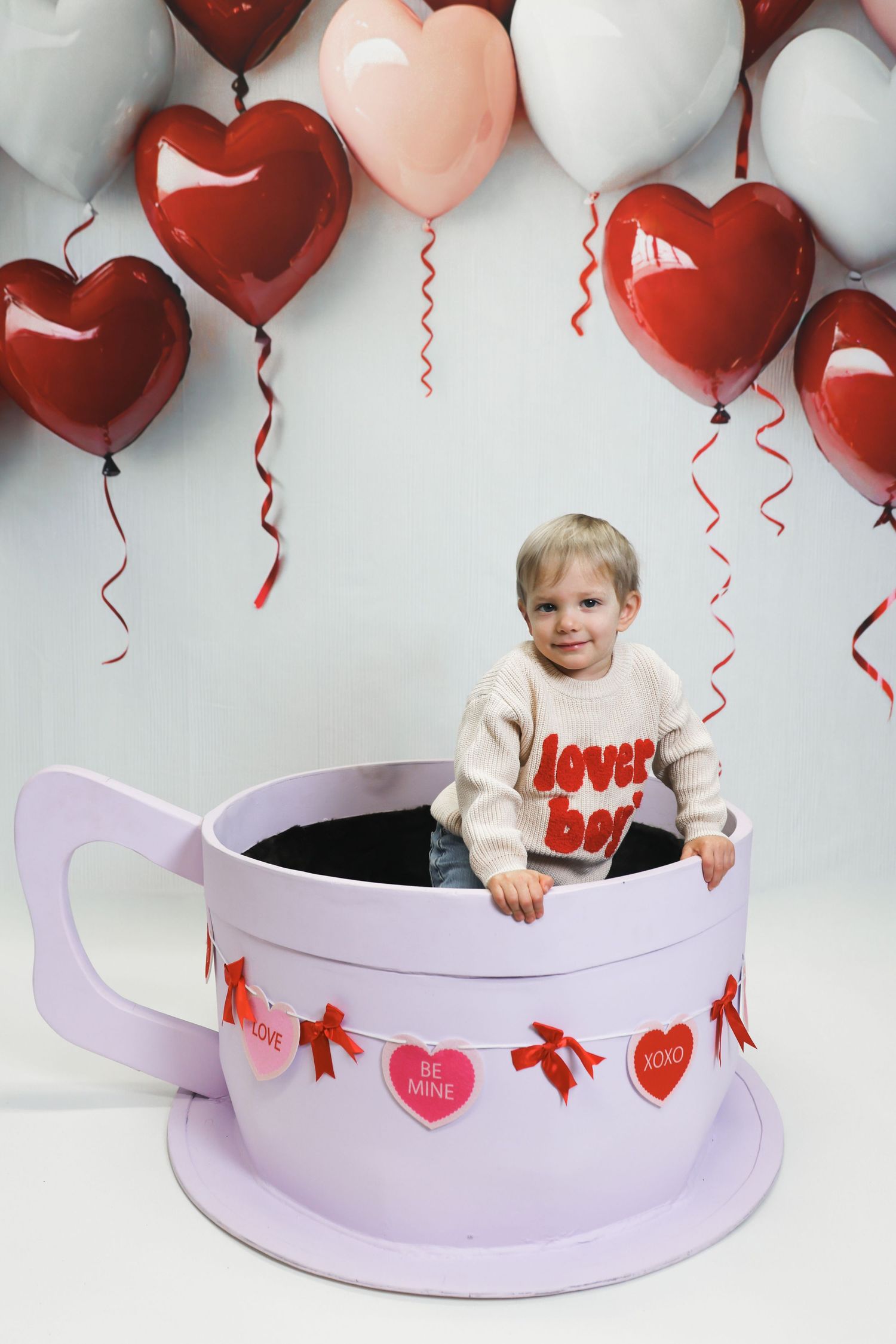 Valentine's day themed photo setup with heart balloons and large decorative purple teacup against white background.