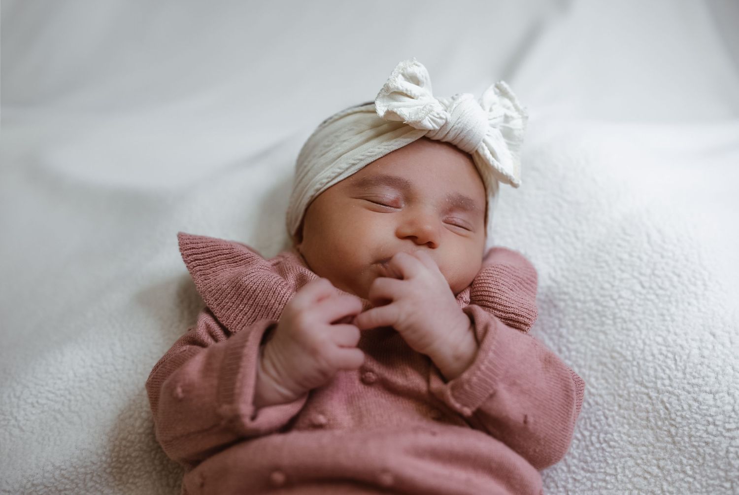 Sleeping baby wearing a pink sweater and white bow headband rests peacefully on white bedding.
