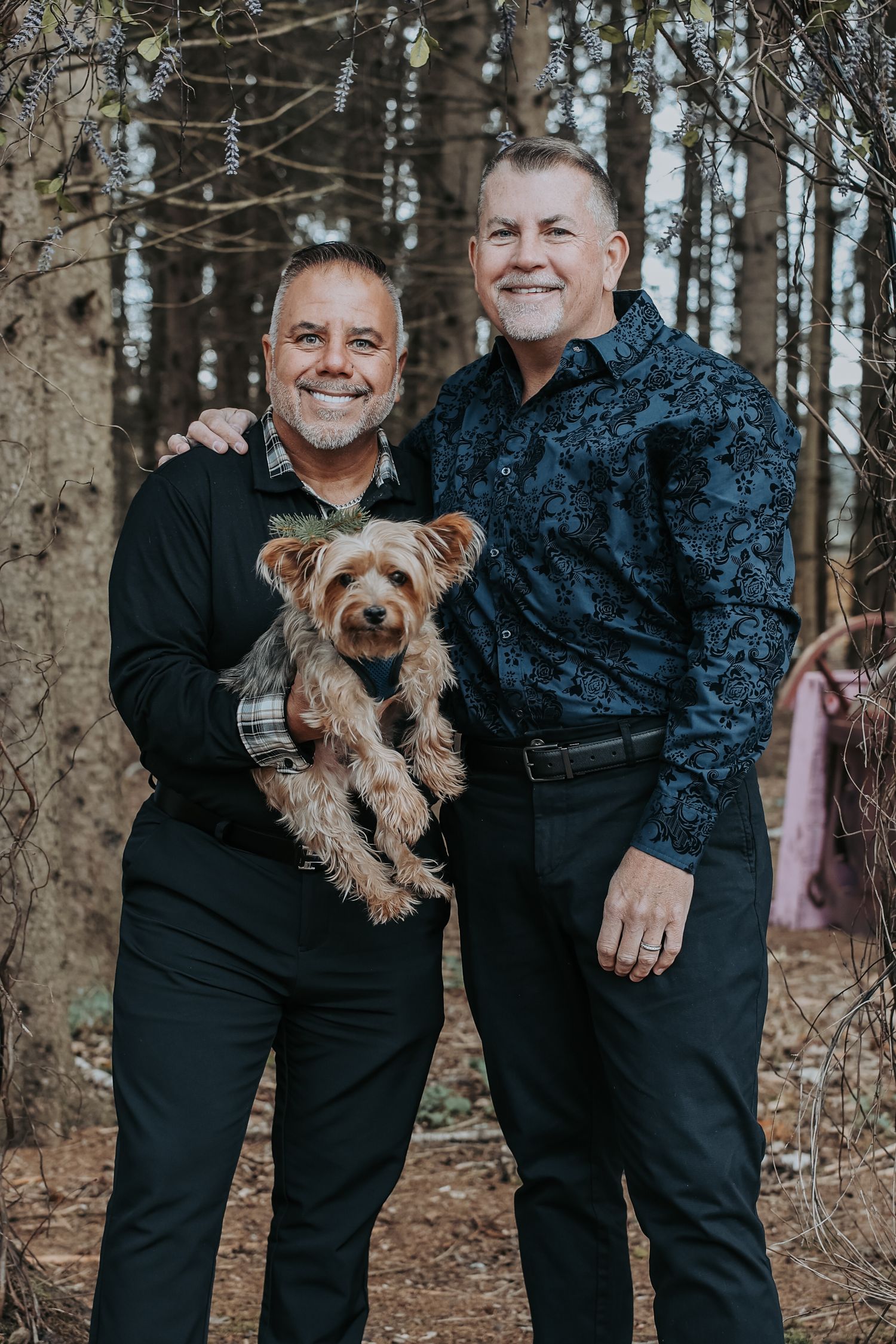 A couple in black outfits poses with their small brown Yorkshire Terrier dog in a forest setting.