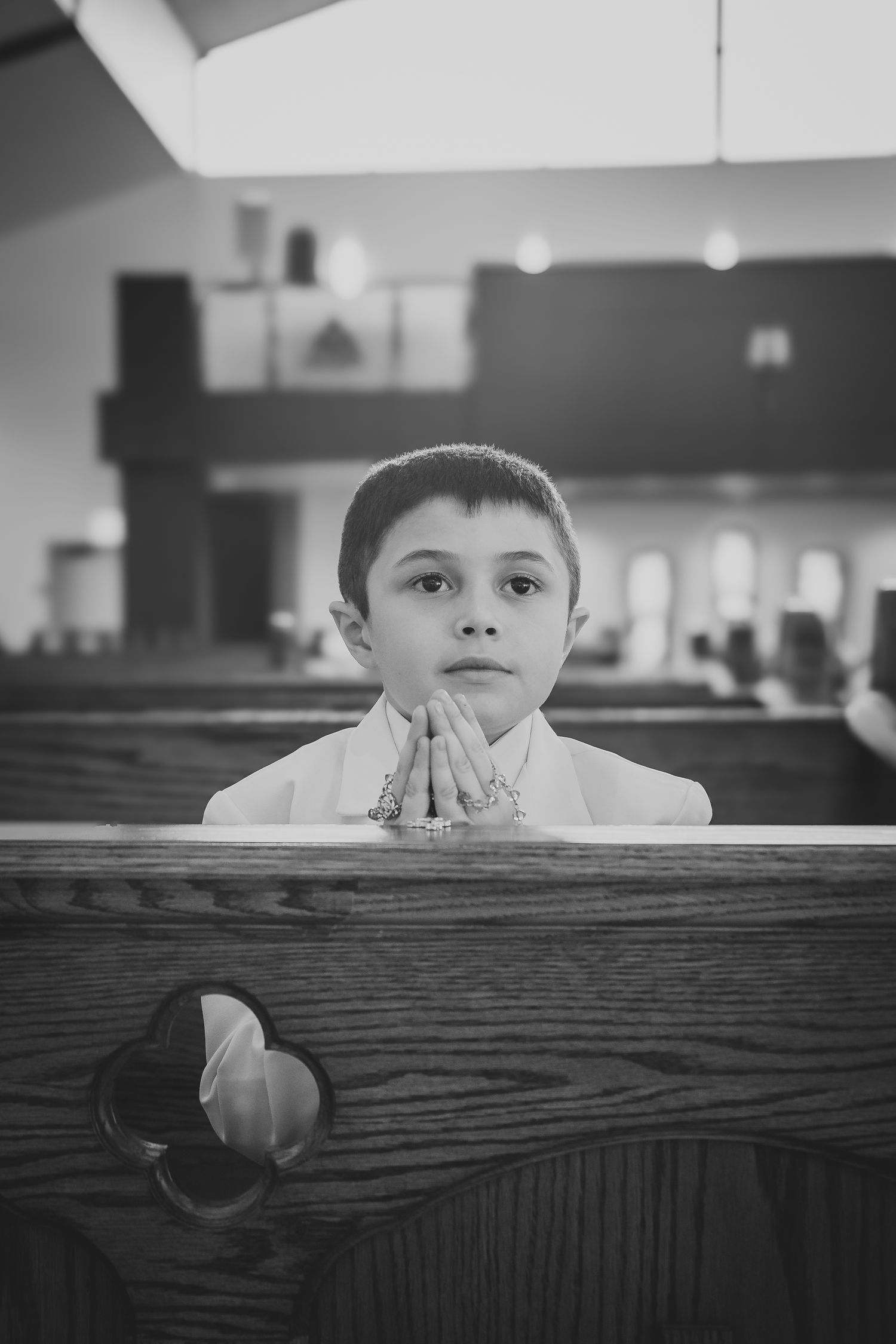Black and white photo of a child praying while kneeling at a wooden church pew.