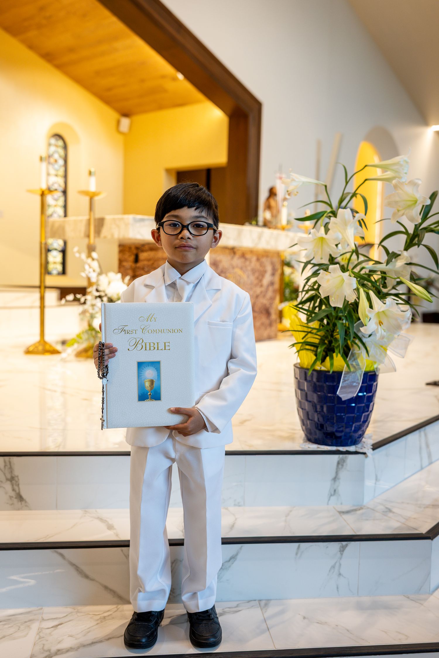 A young student in a white suit holds a religious book while standing in a church interior with flowers.