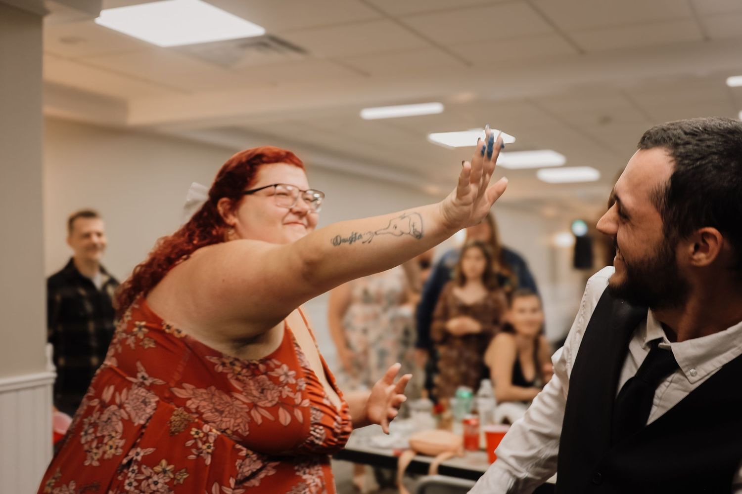 A couple shares a joyful dance move during what appears to be a wedding reception in a bright indoor venue.
