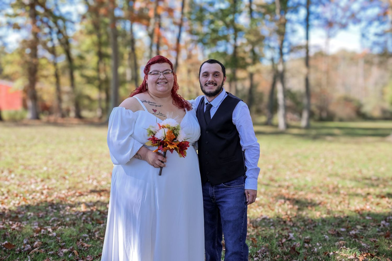 A couple poses together in a park during autumn with fallen leaves scattered on the ground.