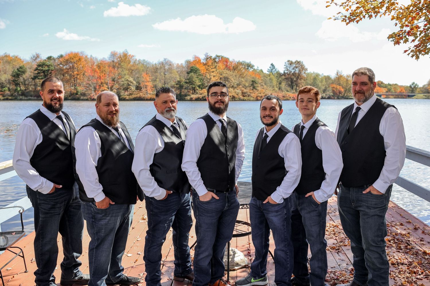 Groomsmen in matching black vests and white shirts pose together by a lake during autumn.