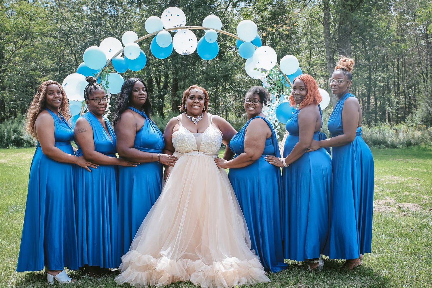 A wedding party poses outdoors with bridesmaids in bright blue dresses and the bride in a light blush gown.