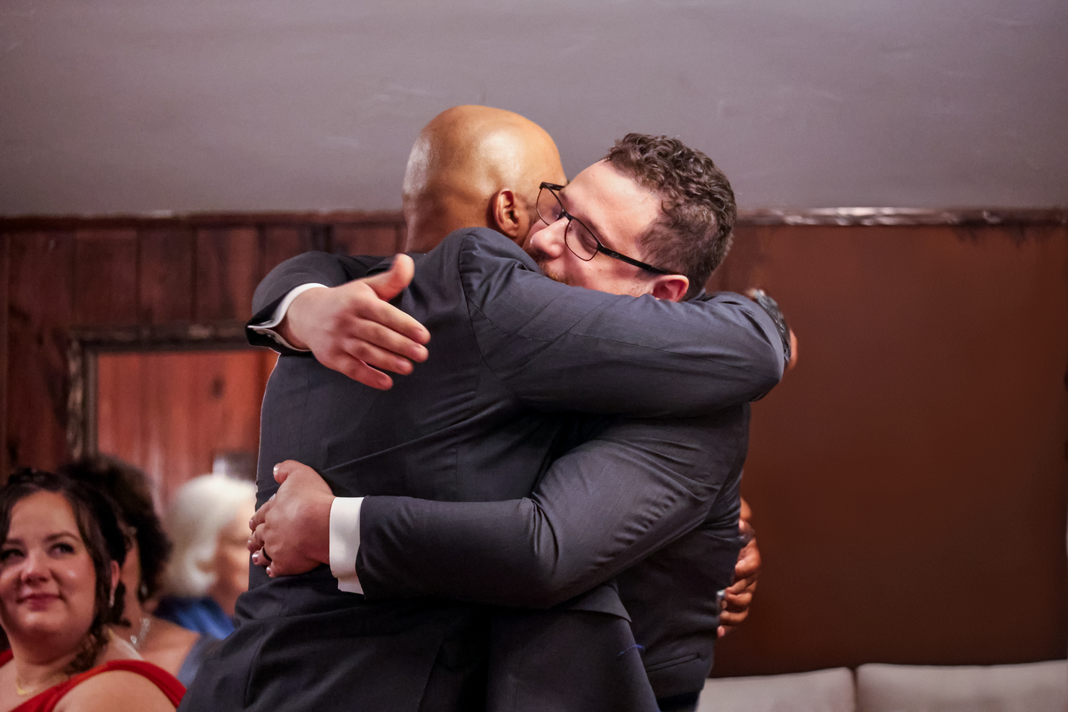 Two people in formal suits share an emotional embrace at an indoor event with onlookers in the background.