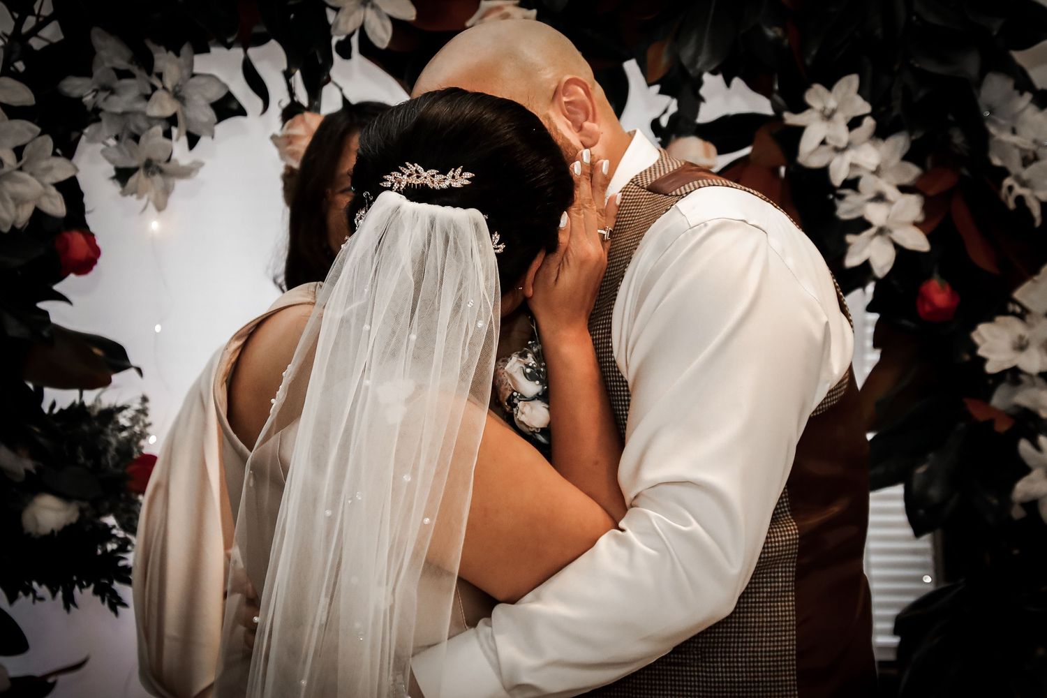 Couple in white attire sharing a romantic kiss during their wedding celebration with floral decor in the background.