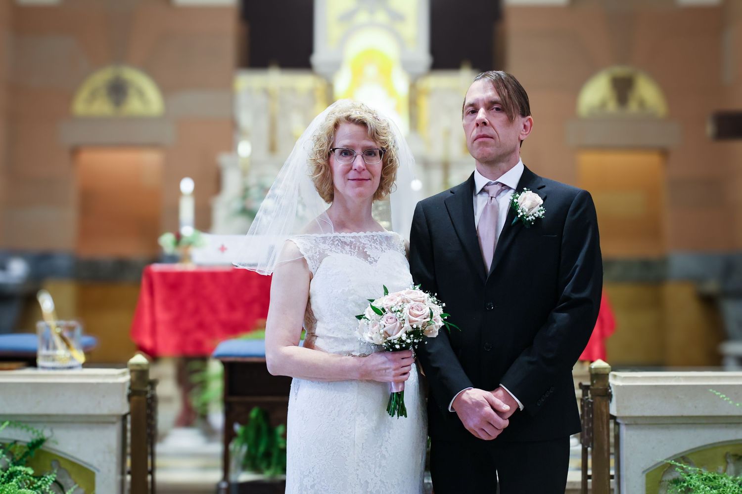 A wedding party walks down a church aisle with white floral decorations and red accents.