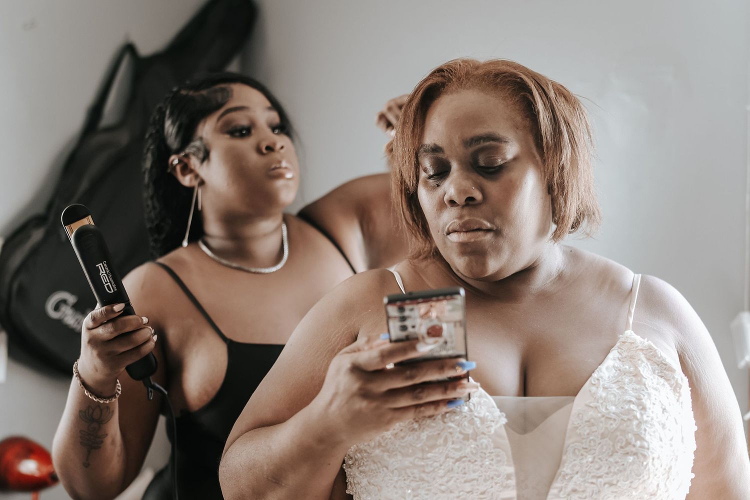 Two women in elegant attire pose together while holding drinks and a phone at a special event.