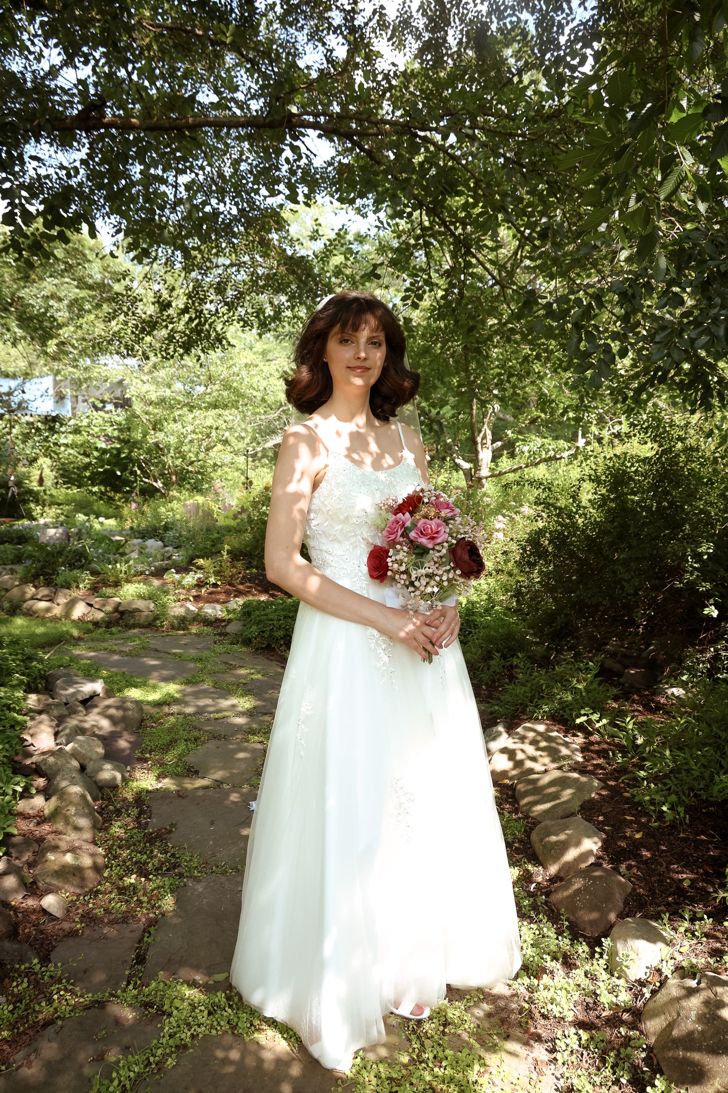 A bride in a white wedding gown holds a deep red bouquet while standing in a sunlit garden path surrounded by greenery.