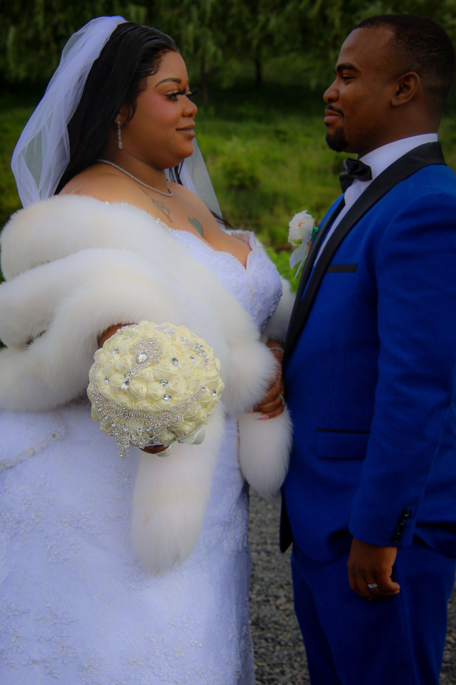A couple in wedding attire, with the bride in a white fur-trimmed dress holding a cream bouquet and groom in royal blue suit.