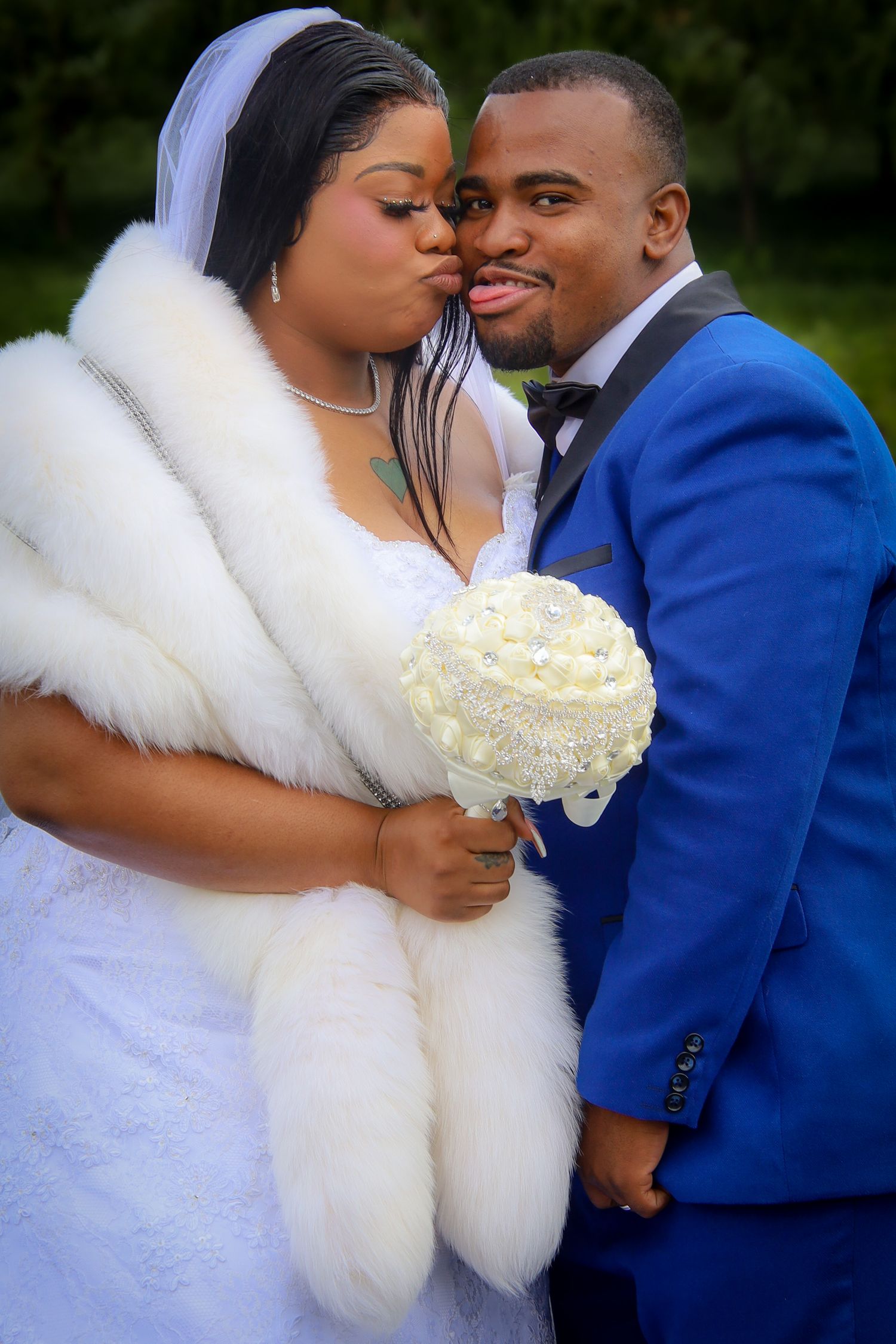 A romantic wedding couple embraces outdoors, with the bride in white lace and fur wrap and groom in royal blue suit.