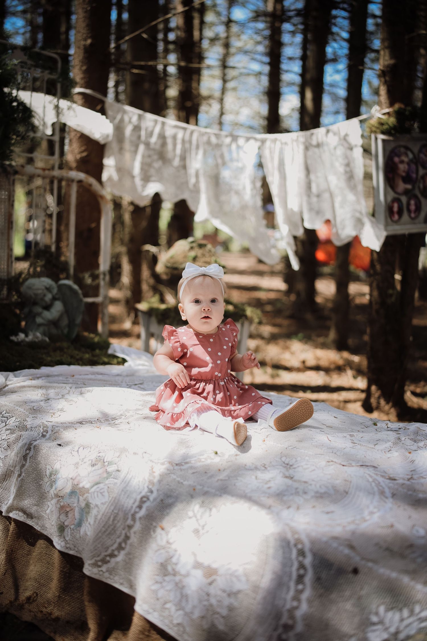 Baby girl in pink polka dot dress sitting on white lace tablecloth in forest with bunting decorations.