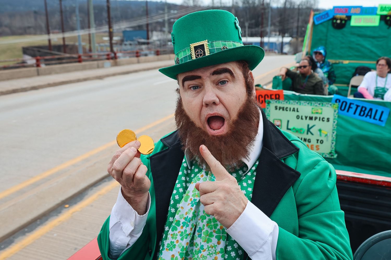 Man dressed as leprechaun in green suit and hat holds gold coin at St. Patrick's Day parade.