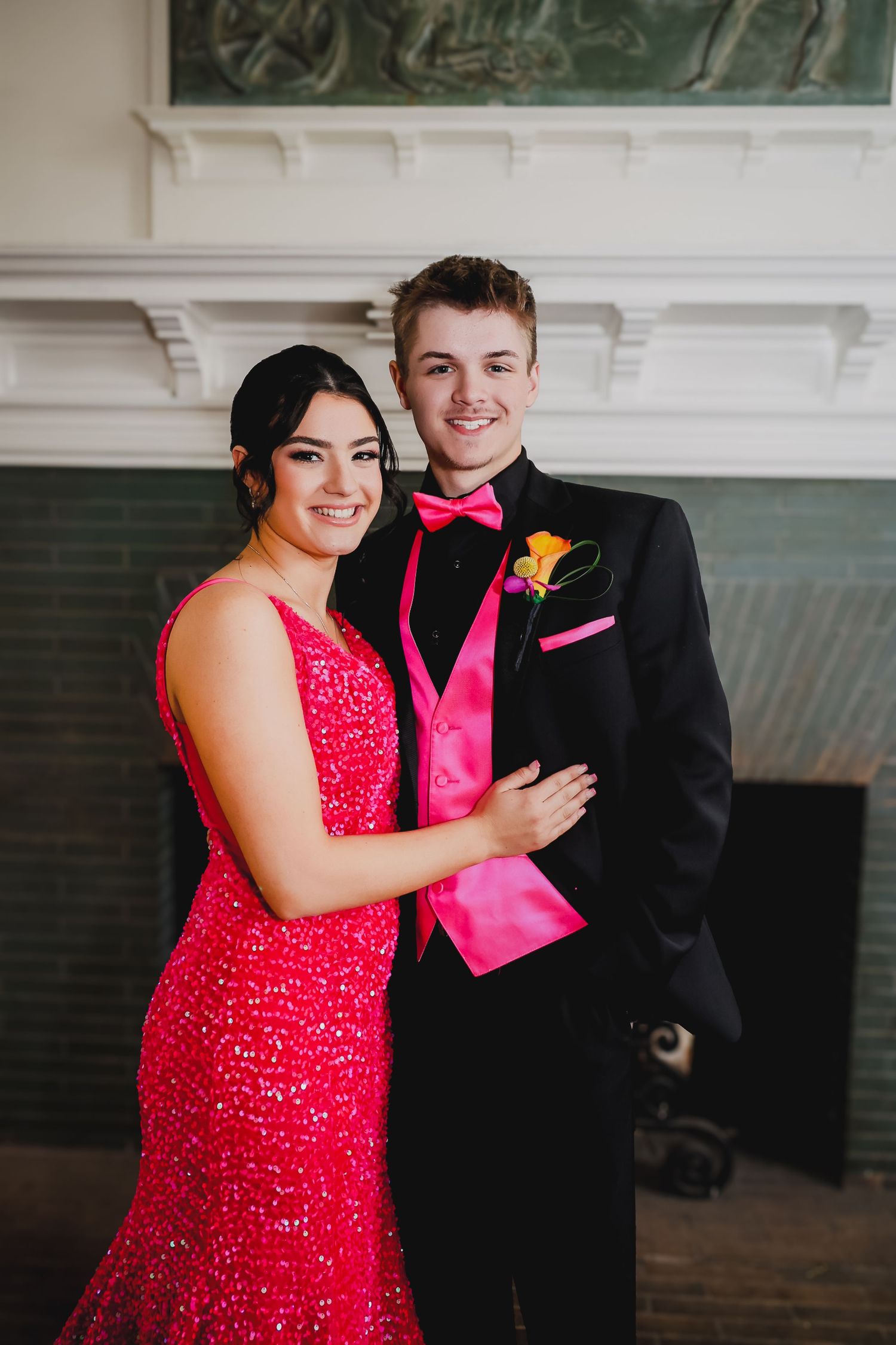 A couple dressed in prom attire, woman in red sequin gown, man in black tuxedo with pink vest and bow tie.
