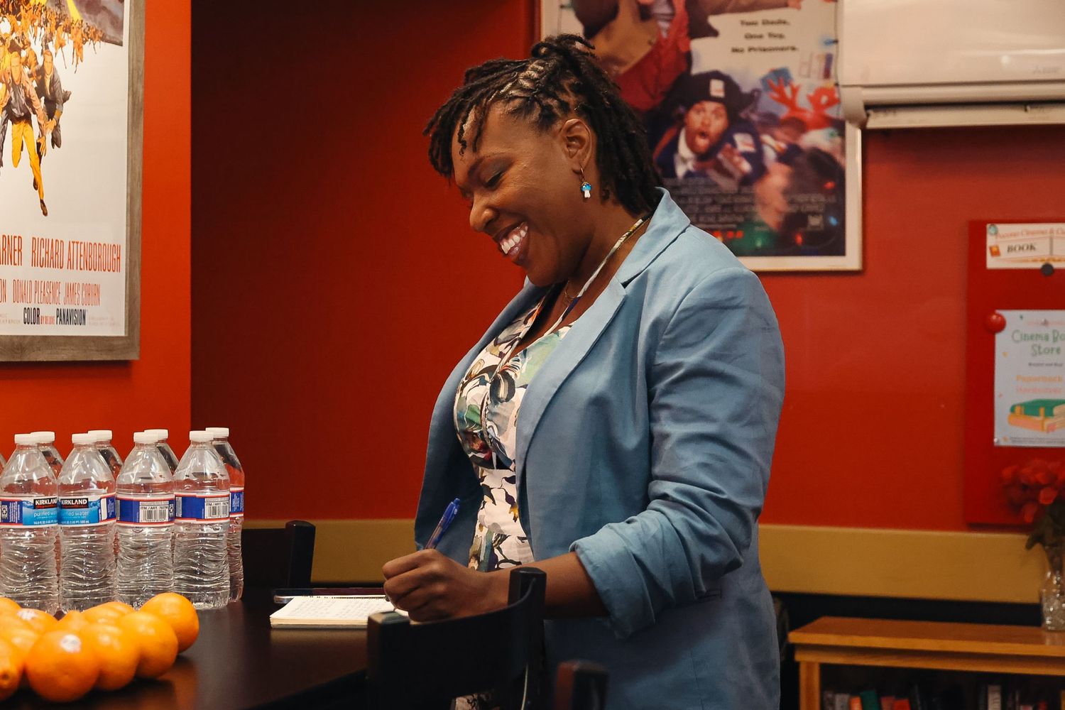 A smiling woman in a blue blazer stands at a counter with water bottles and oranges in a bright office.