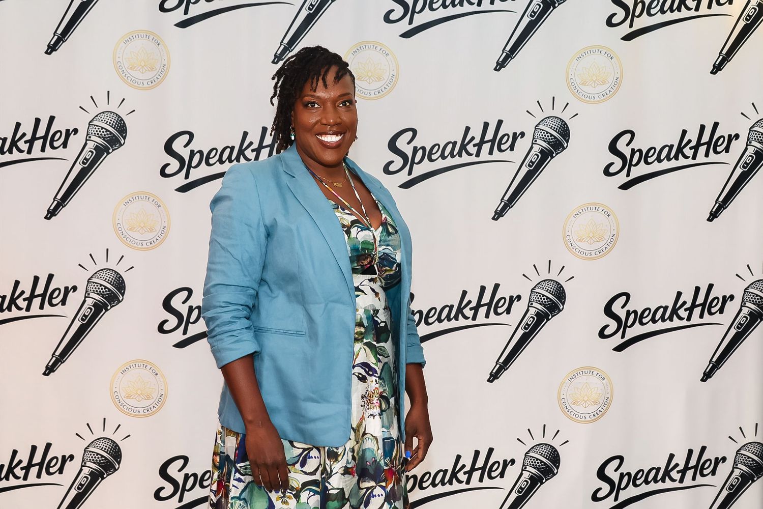 Woman in floral dress and blue blazer posing at SpeakHer event backdrop with microphone logos.