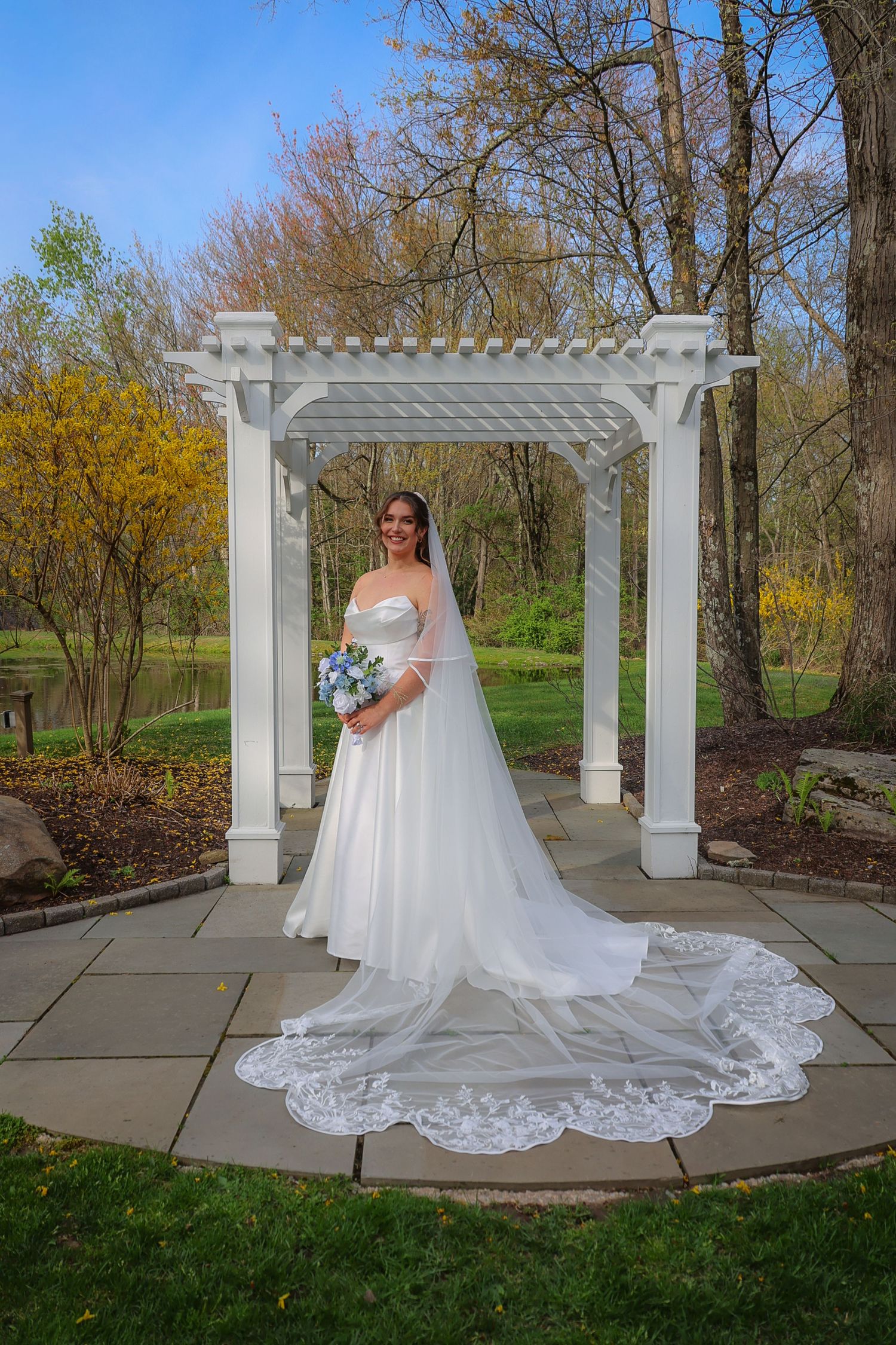 Bride in a white lace-trimmed gown and veil holding a bouquet, standing under a white pergola in a garden.