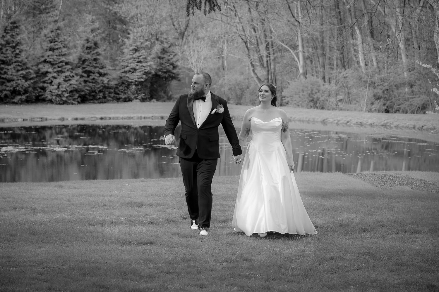 Black and white photo of a bride and groom holding hands walking across a lawn near a pond on their wedding day.