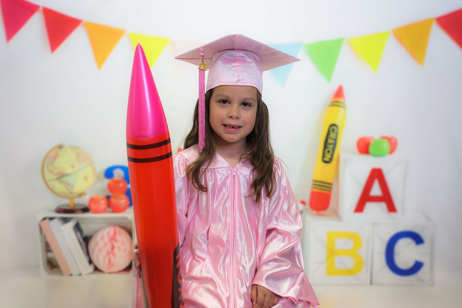 Young girl in pink graduation cap and gown holding oversized crayon with ABC blocks and school supplies in background.