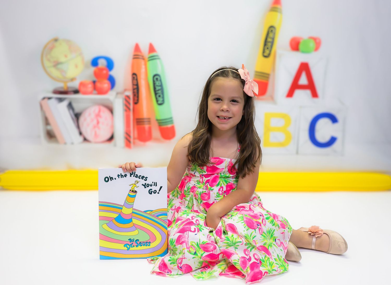 Young girl in floral dress sits with Dr. Seuss book, surrounded by colorful ABC letters and crayons backdrop.