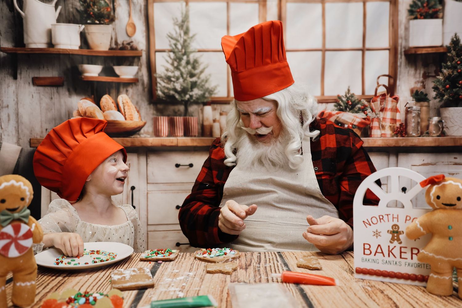 Child and Santa Claus in red chef hats decorating Christmas cookies together in a festive holiday kitchen.