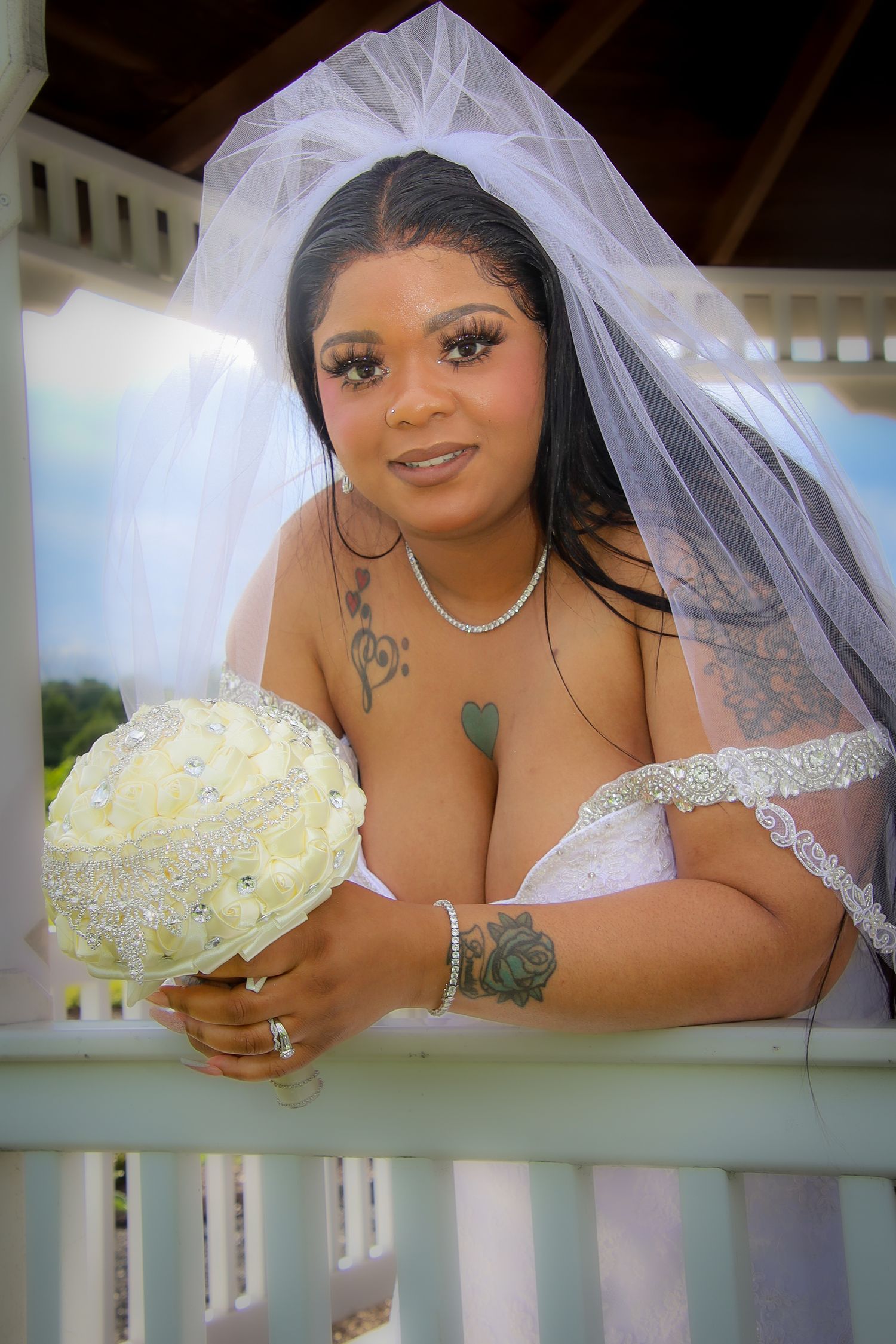 A bride in a white wedding dress and veil poses while holding a white rose bouquet against a railing.