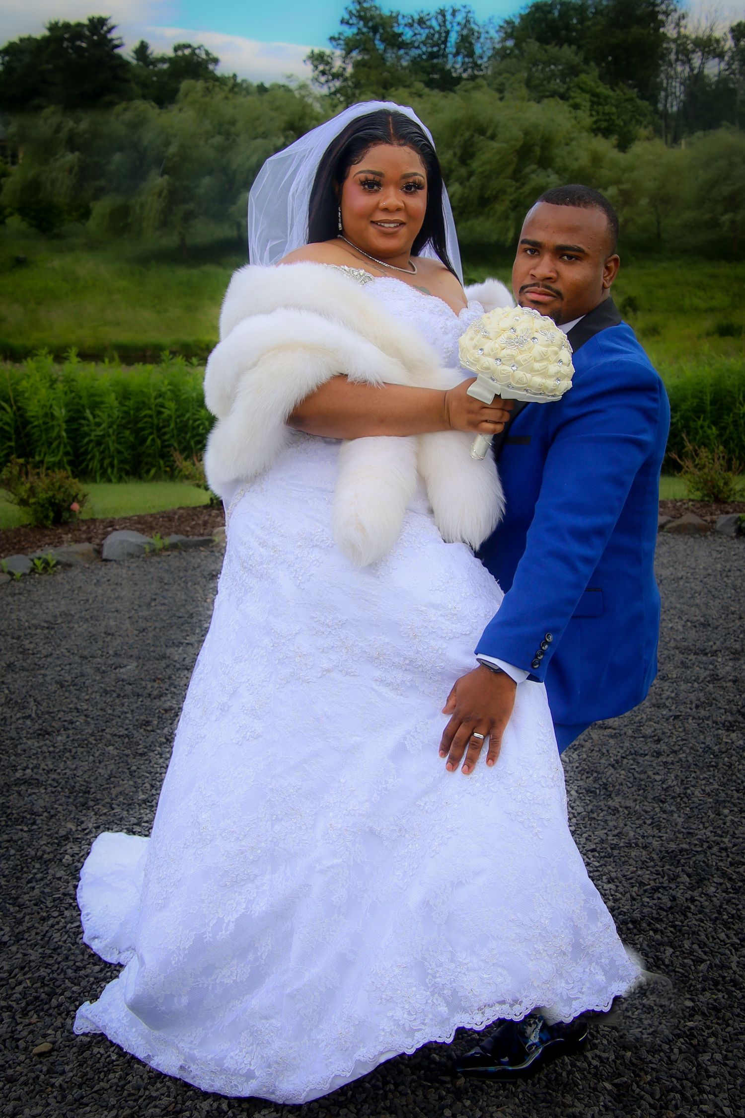 A couple in wedding attire shares a romantic moment outdoors, with the bride in a white lace gown and groom in a blue suit.