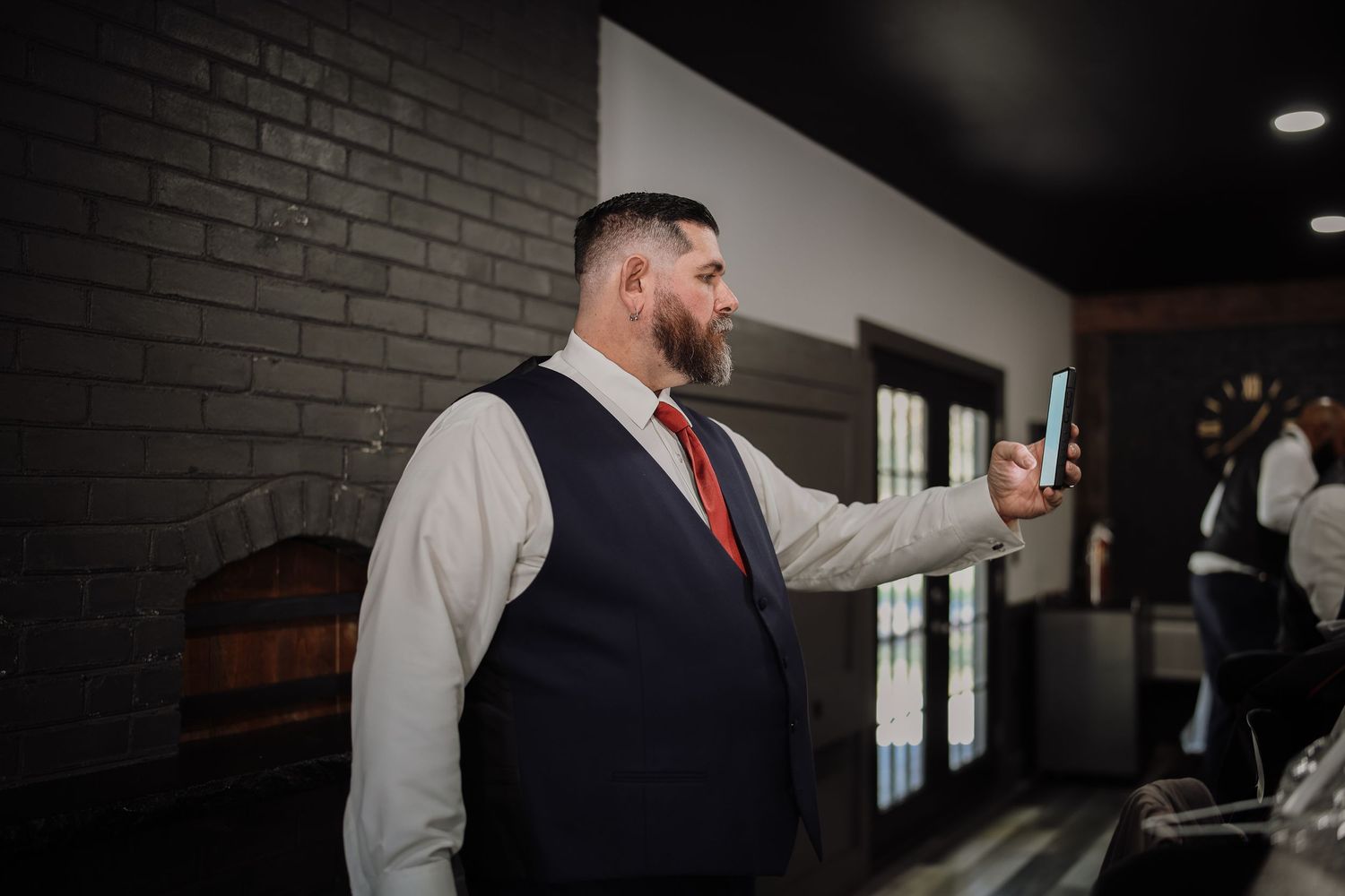 A person in a navy vest and red tie adjusts a tie while standing in front of a dark brick wall.