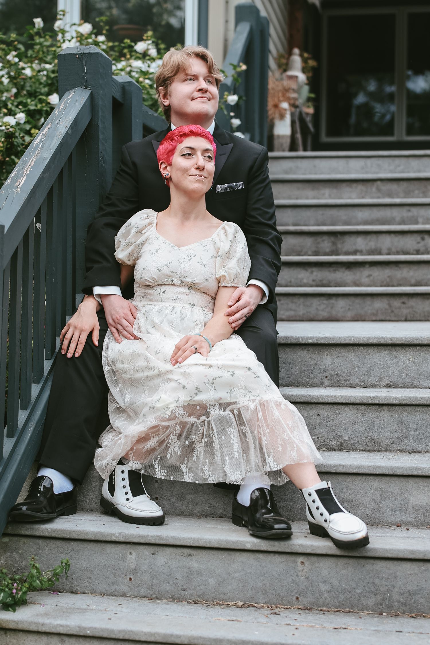 A couple sits on outdoor stairs, with one wearing a vintage white lace dress and the other in black, both wearing sneakers.