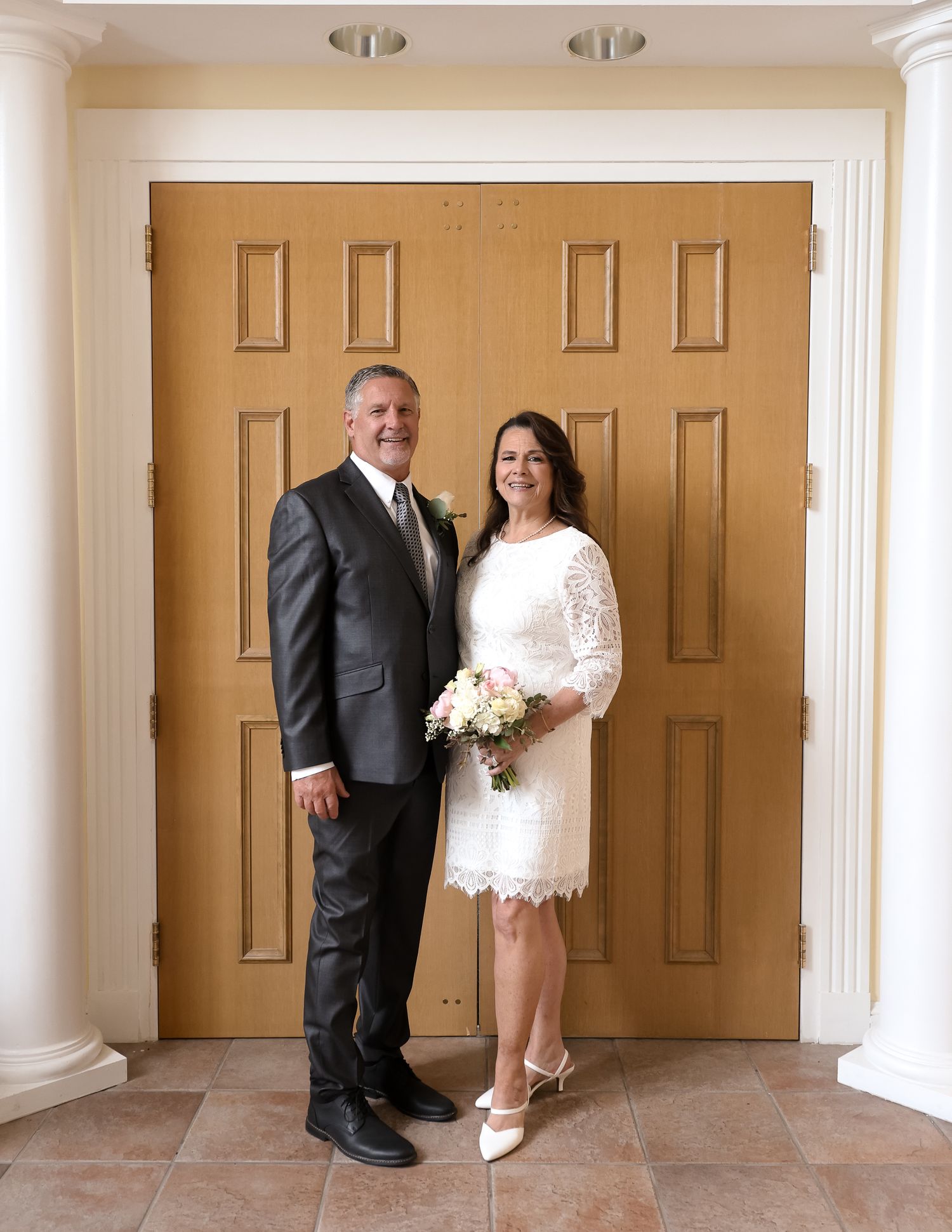 A couple poses in front of a wooden door wearing formal attire, with the bride holding a white bouquet.